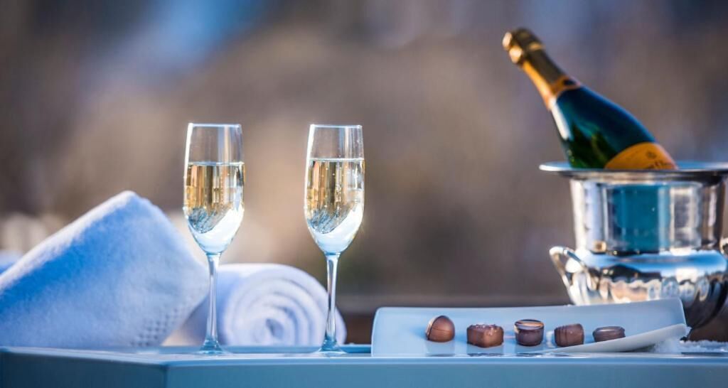 Two glasses of white wine, a bottle of champagne, and a tray with nuts on a table with a towel and a silver container.