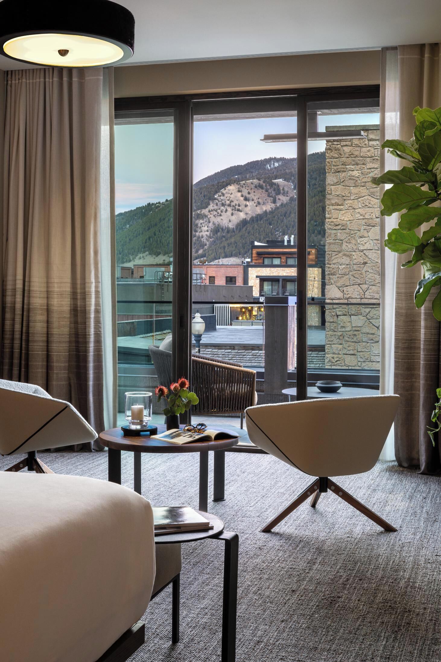 A hotel room with a large window, a bed, a chair, a table with a candle and flowers, and a potted plant.