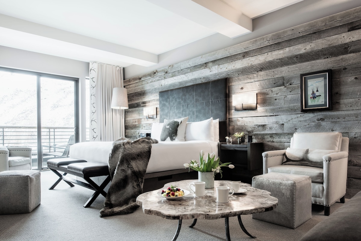 A modern hotel room with a large bed, a sitting area with a coffee table, and a view of the outdoors.