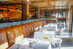 Een restaurant met een lange, bruine leren zitbank, witte tafels met gedekte servies en een bar op de achtergrond.