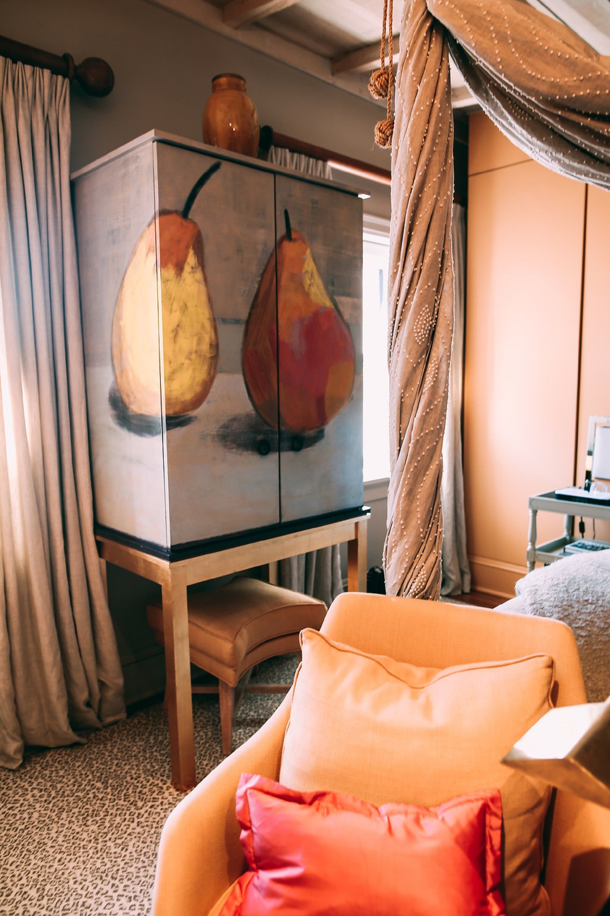 A room with a wooden cabinet featuring pear paintings, a beige armchair with a red pillow, and a small table with a lamp.