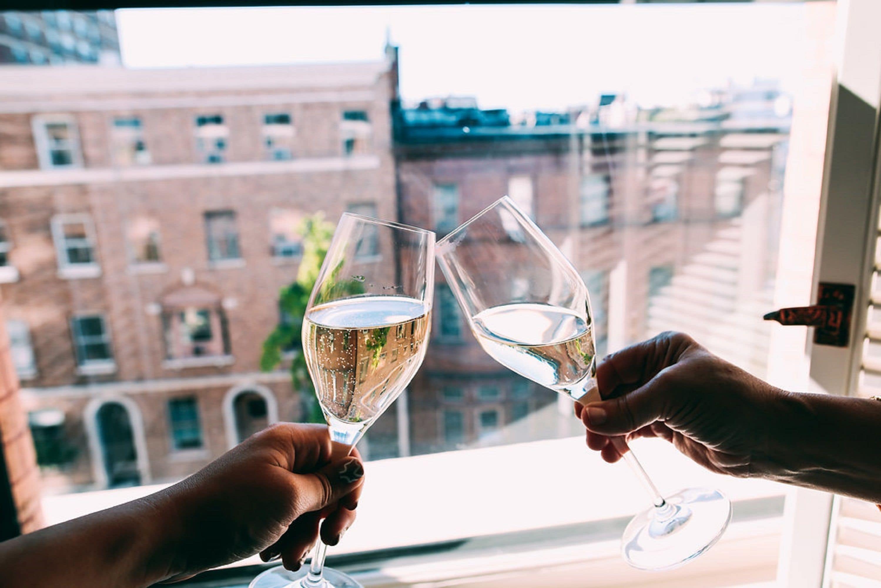 Two hands holding wine glasses with a cityscape view through a window.
