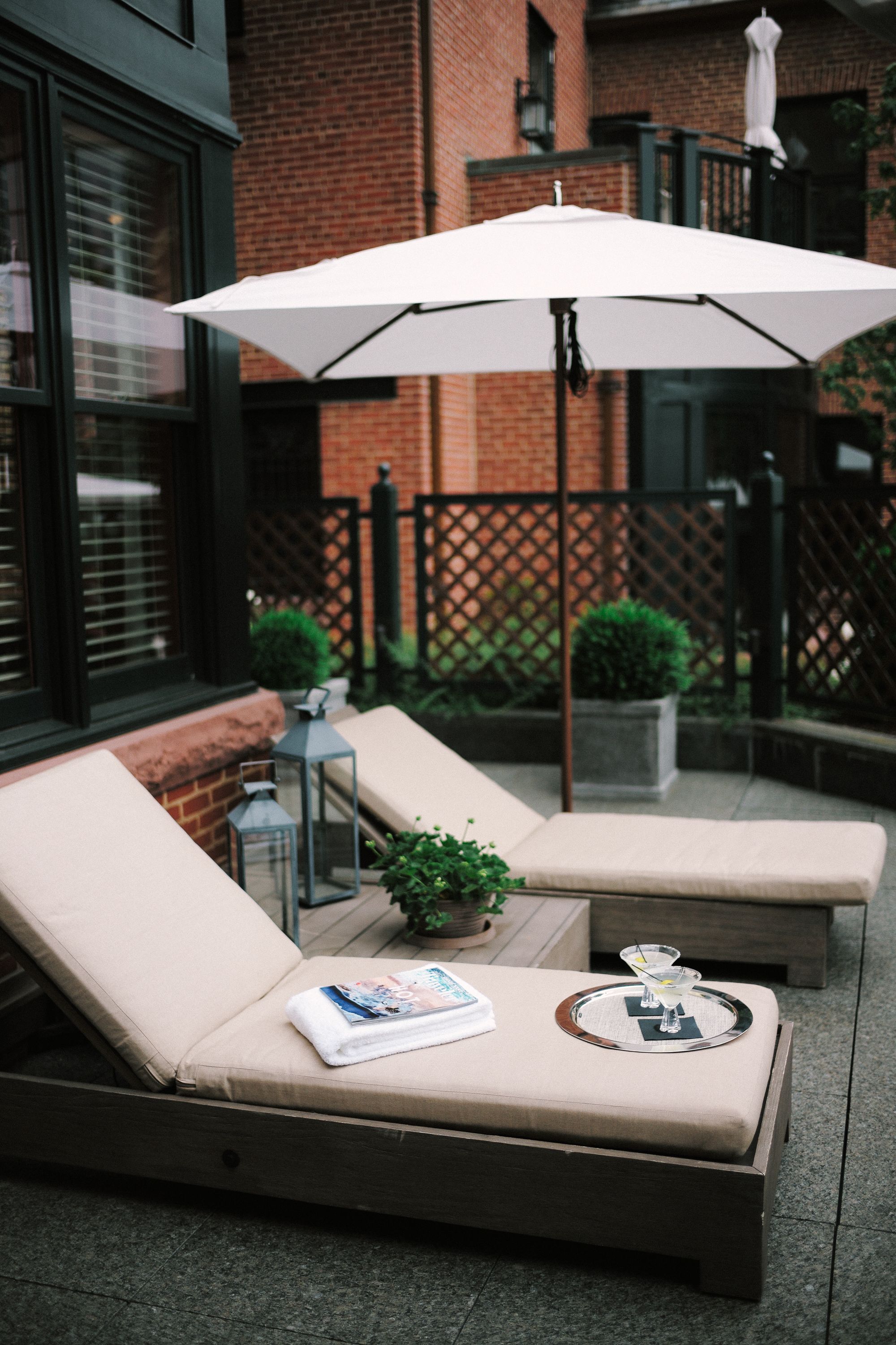 Un patio con dos sillones de descanso, una sombrilla blanca y una mesa pequeña con una revista y un vaso.