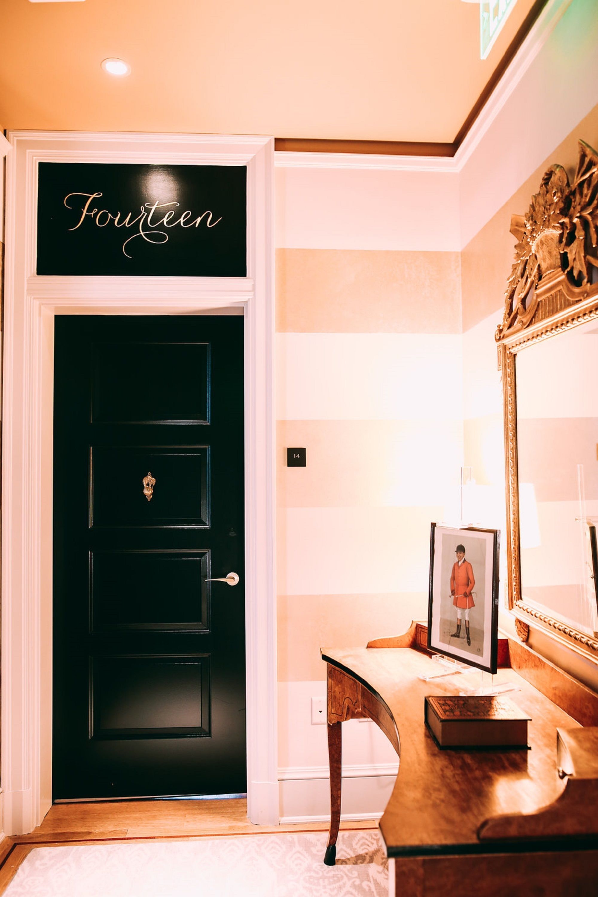 A black door with a sign reading "Fourteen" above it, a wooden desk with a framed picture, and a large mirror on the wall.