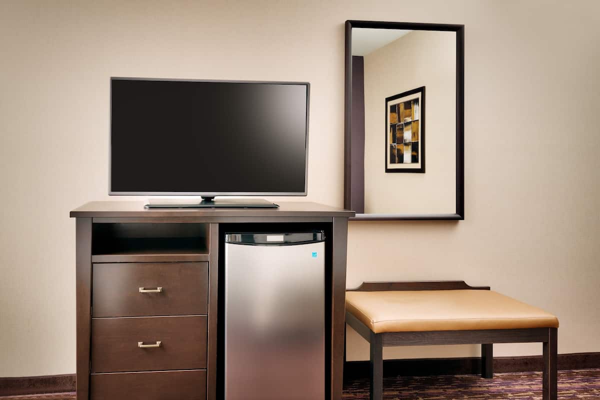 A hotel room with a desk, a flat-screen TV, a refrigerator, a bench, and a mirror reflecting a framed picture.