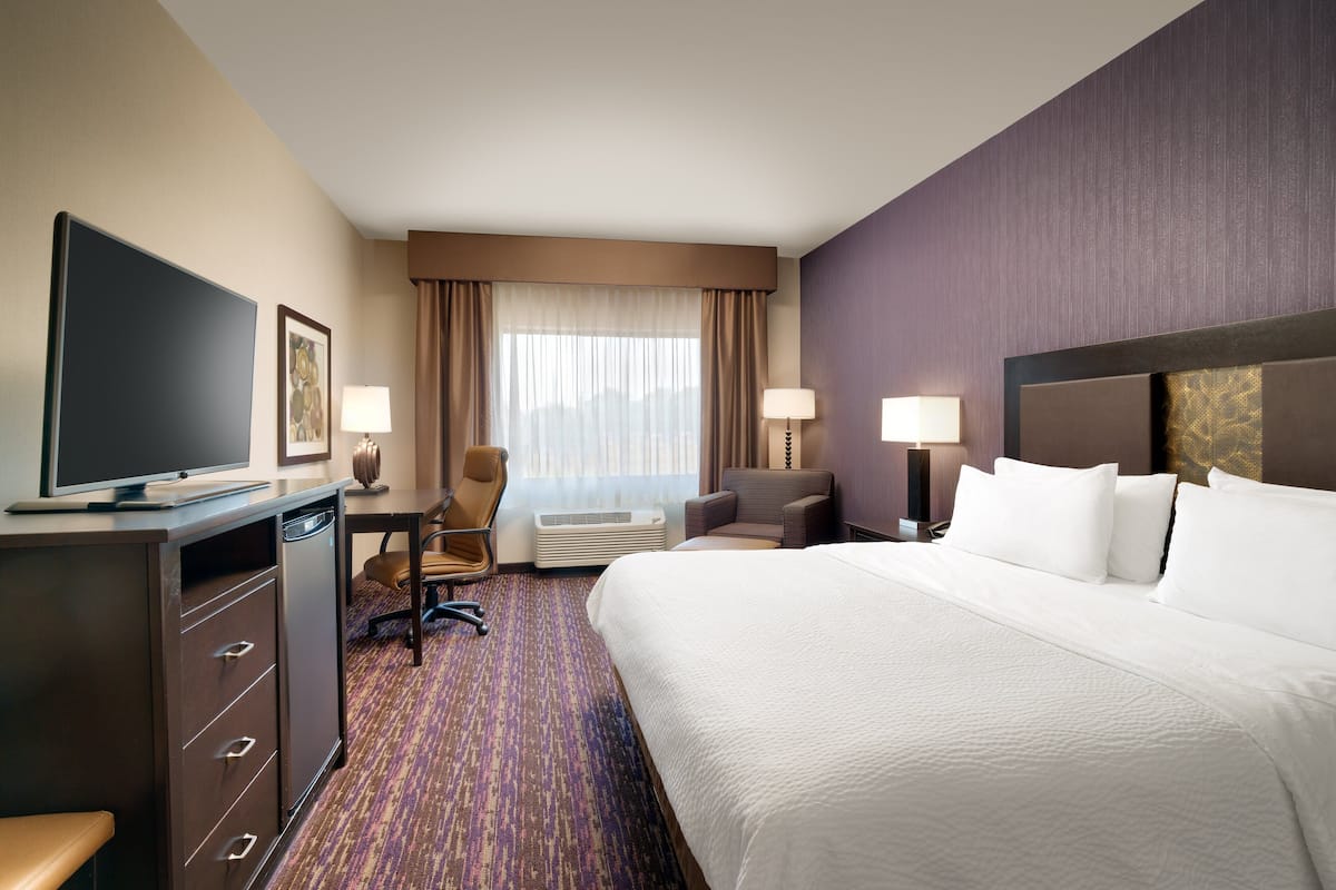 A hotel room with a large bed, a desk with a computer, a chair, a television, and a window with curtains.