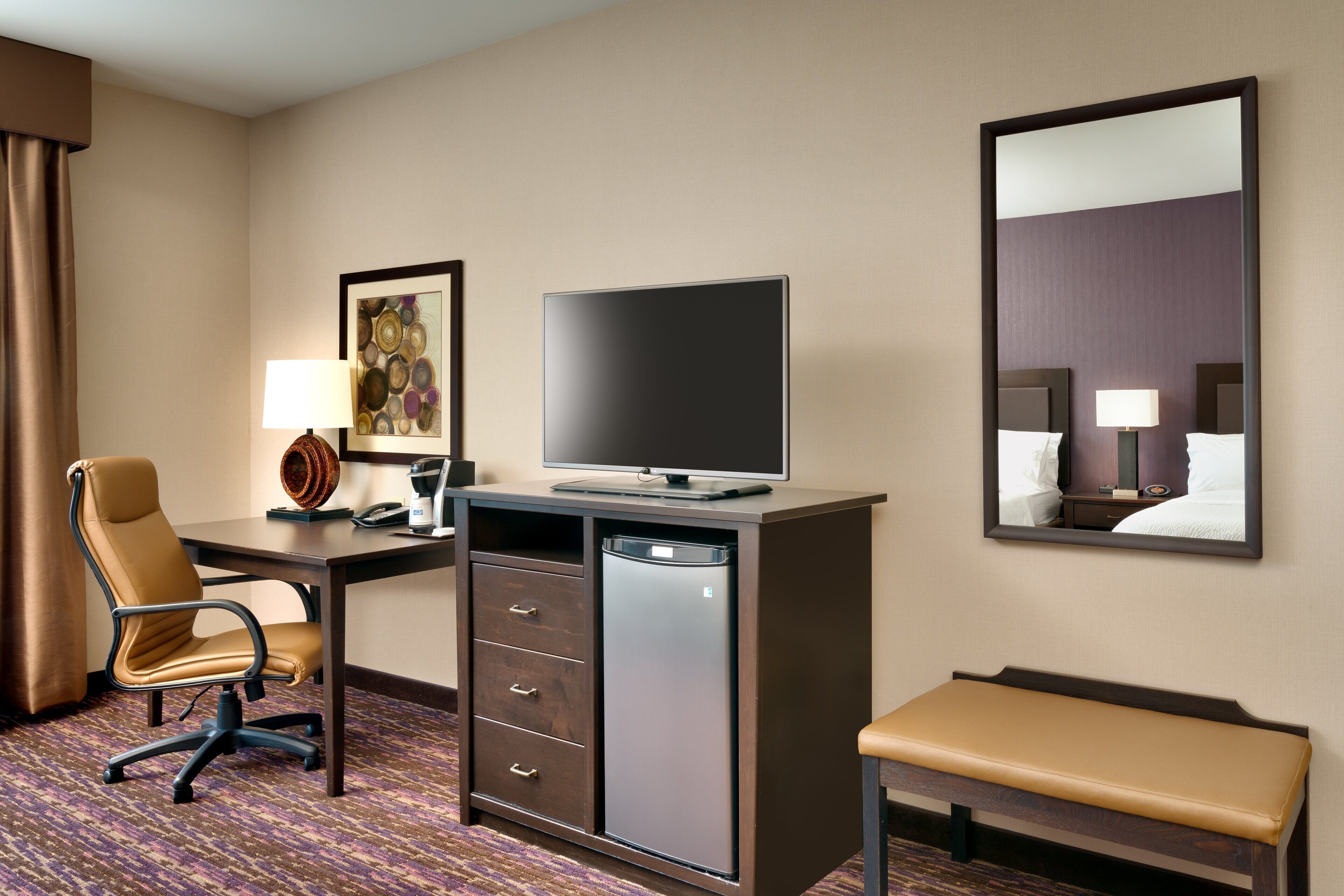 A hotel room with a desk, chair, television, and a bench.