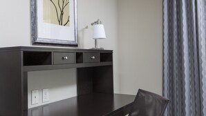 Ein dunkles Holzschreibtisch mit einem Stuhl, ein gerahmtes Bild an der Wand, eine Lampe und ein gemustertes Vorhang.
