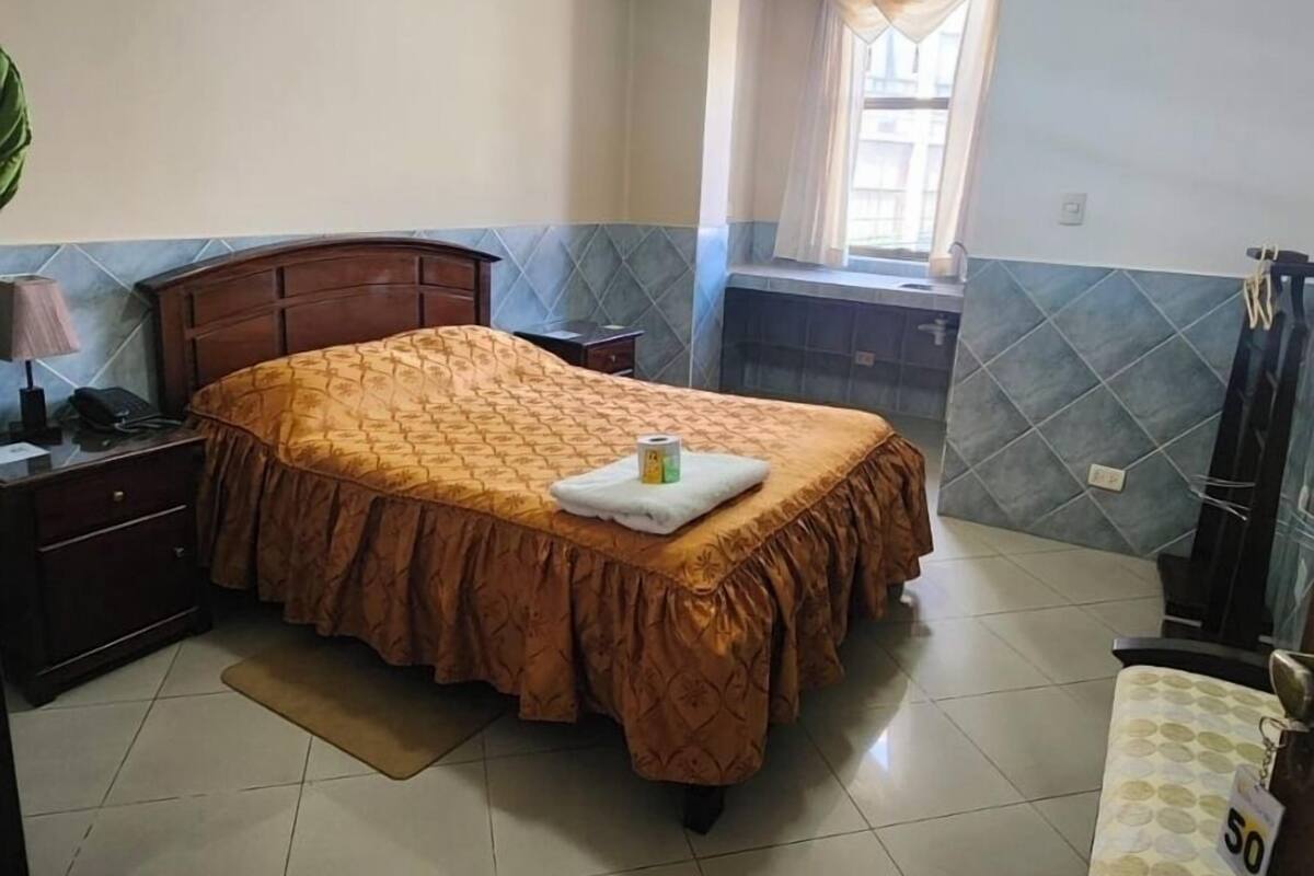 Ein Hotelzimmer mit Bett, Nachttisch, Schreibtisch, einem Fenster mit Vorhängen und einem im Hintergrund sichtbaren Badezimmer.