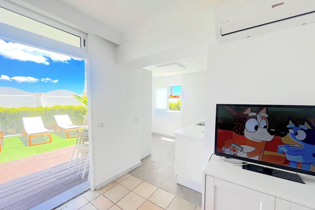 Ein heller, moderner Wohnraum mit einem großen Fernseher, einer Glasschiebetür, die auf eine Terrasse mit Liegestühlen führt, und einer Küchenzeile.