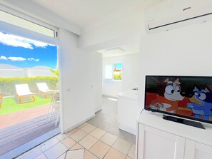 A bright, modern living room with a large TV, a sliding glass door leading to a patio with sun loungers, and a kitchen area. - Apartamentos Dunaoasis Maspalomas (San Bartolomé de Tirajana)