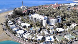 An aerial view of a coastal resort with a beachfront hotel, palm trees, and a lighthouse in the background.