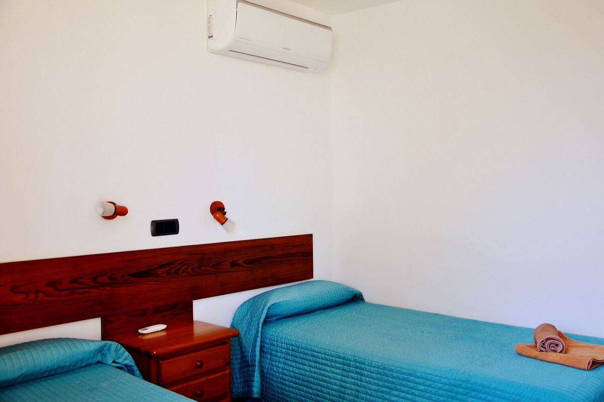 Ein Zimmer mit einem hölzernen Kopfende, einem Bett mit blauer Steppdecke, einem Nachttisch aus Holz, einer Fernbedienung und einer Klimaanlage.