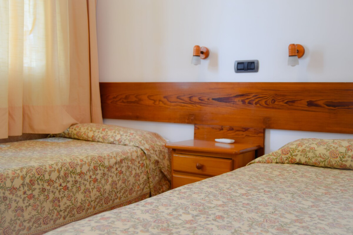 Une chambre d’hôtel avec deux lits simples, une tête de lit en bois, une table de chevet avec une lampe et une petite table de nuit avec un interrupteur.