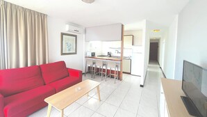 A compact living space with a red sofa, a wooden coffee table, a kitchen area with a microwave and a refrigerator, and a television on a stand.