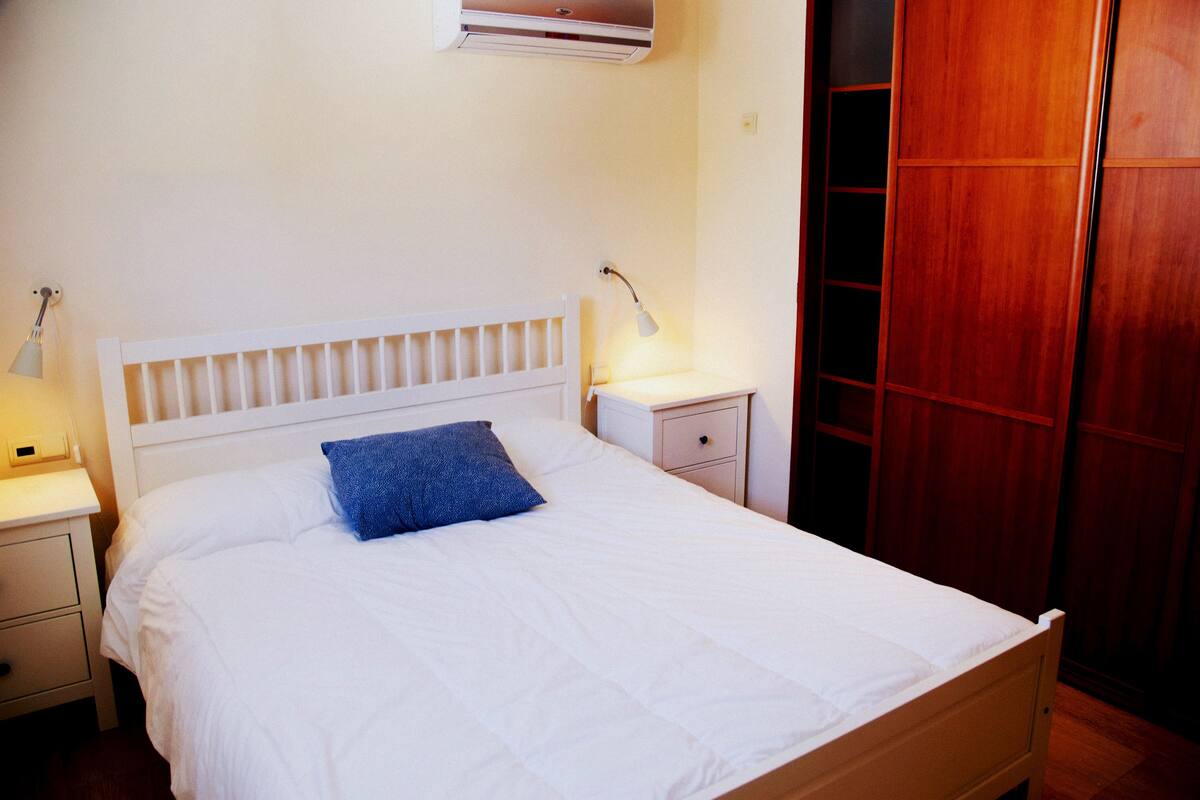 Ein ordentlich bezogenes Bett mit weißem Kopfteil und blauem Kopfkissen, ein Nachttisch mit Lampe sowie ein Holzschrank.