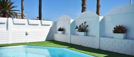 A swimming pool with white brick walls and green artificial grass, surrounded by palm trees and white planters with red flowers.