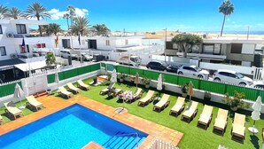 A pool area with sun loungers and a swimming pool, surrounded by a white fence and greenery.