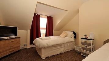 Un dormitorio con cama, televisión sobre una cómoda de madera, ventana con cortinas rojas y mesita de noche blanca con lámpara.