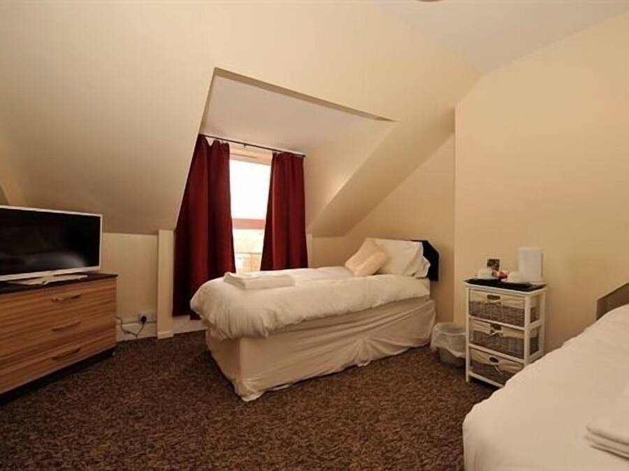 Un dormitorio con cama, televisión sobre una cómoda de madera, ventana con cortinas rojas y mesita de noche blanca con lámpara.
