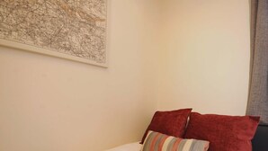 Una cama bien hecha con un almohadón a rayas y dos almohadas rojas, un mapa en la pared y una ventana con cortina.