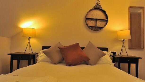Una habitación de hotel con una cama, dos mesitas de noche con lámparas, un reloj de pared redondo y una puerta.