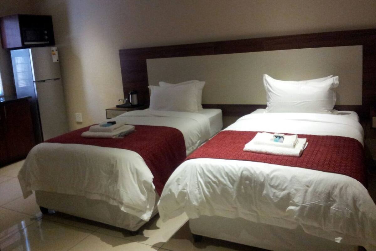 A hotel room with two beds, each with white linens and red bedspreads. There is a wooden headboard, a small table with a lamp, and a refrigerator on the left side.