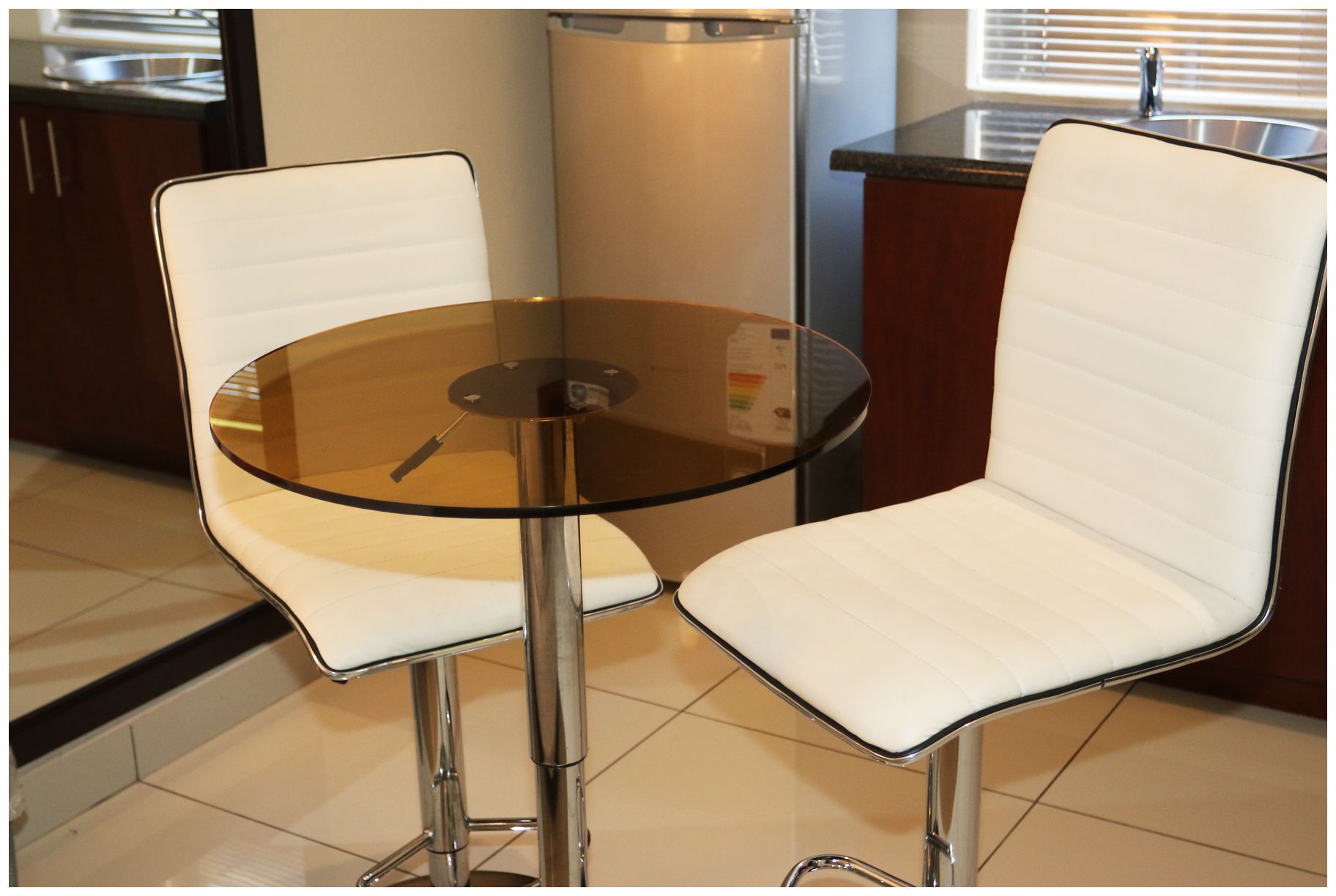 A small, round glass table with a metal base, flanked by two white cushioned chairs with metal legs.
