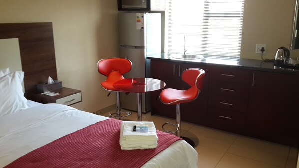 A hotel room with a bed, a small table with two red chairs, a refrigerator, a sink, and a window with blinds.