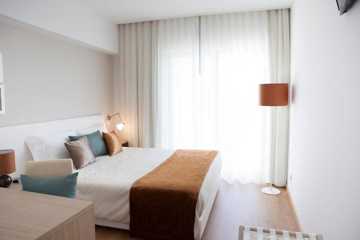 A hotel room with a bed, bedside table, lamp, and a view of the outdoors.