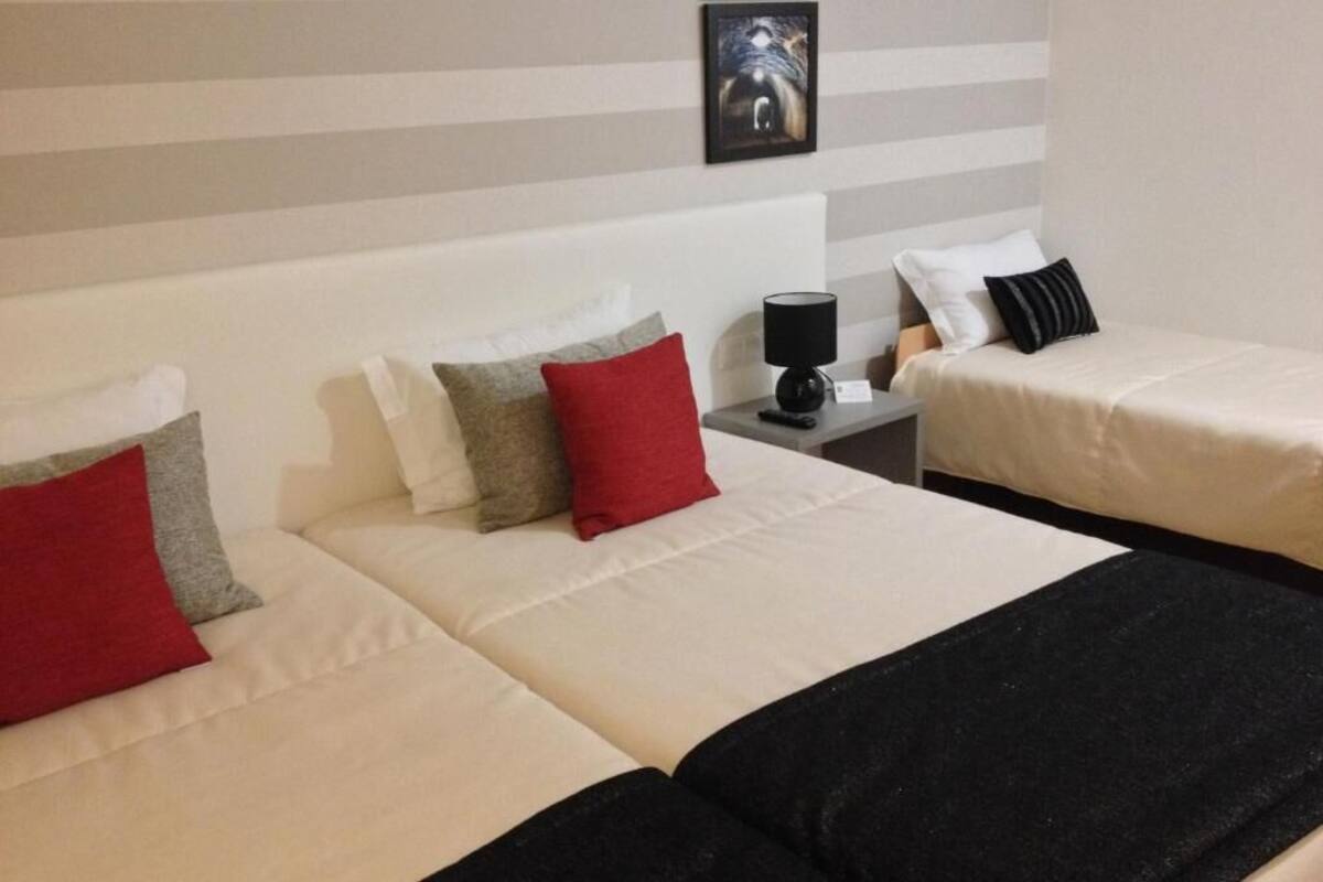 A hotel room with two beds, each with a white mattress and a black blanket. There are pillows in various colors on the beds. A lamp is on the nightstand beside one bed. The wall has a striped pattern and a framed picture above the beds.