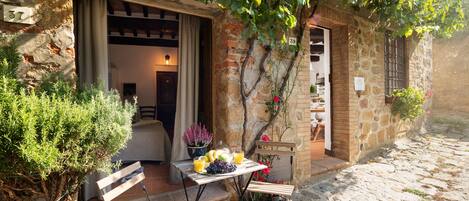 A rustic stone building with a wooden table and chairs outside, a potted plant, and a sign indicating a wine cellar.