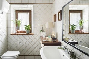 A bathroom with a toilet, a sink, and a bathtub. There are two windows with plants on the windowsills. - In Toscana Affittacamere (Pienza)