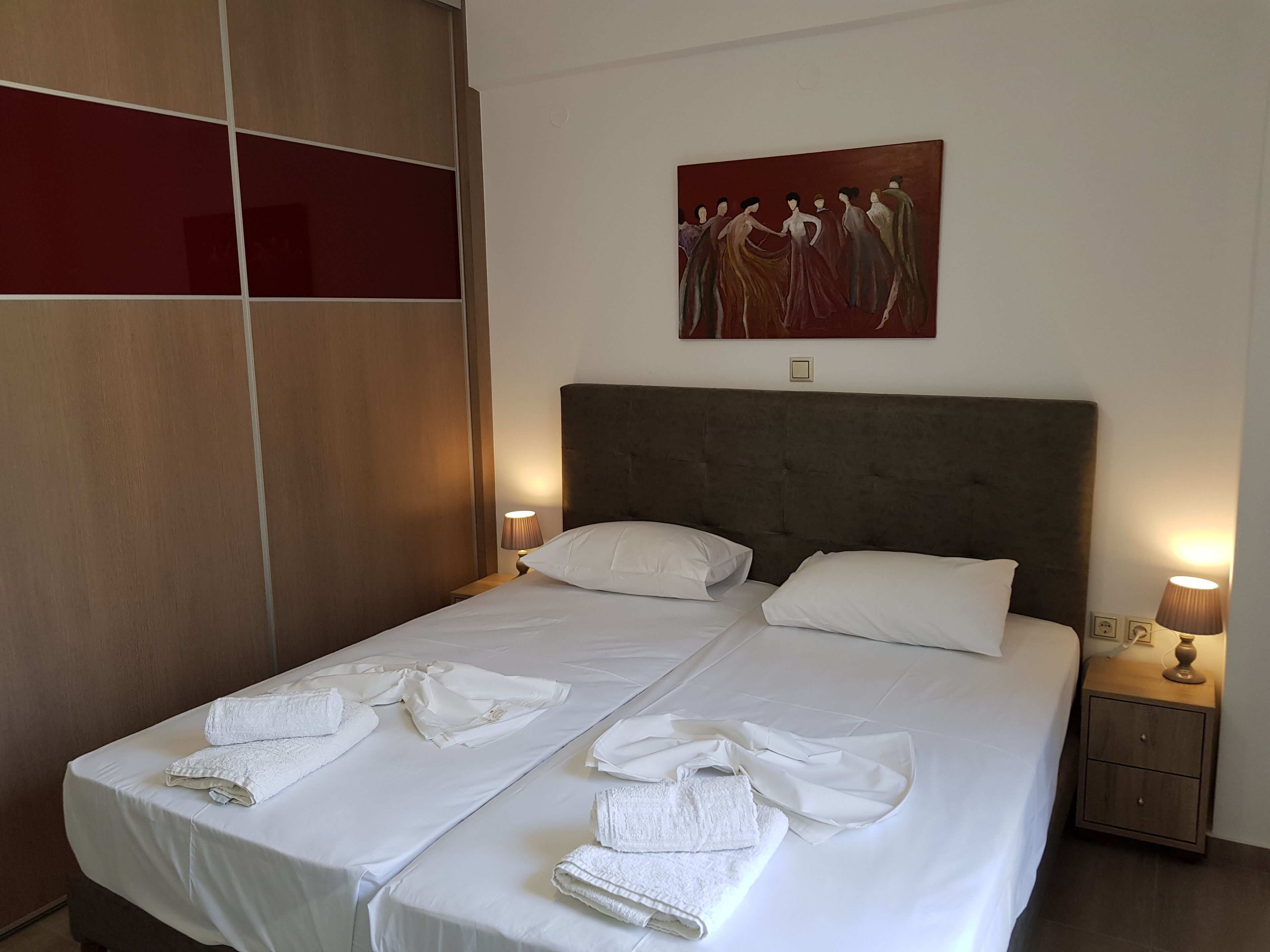 A double bed with white linens and towels, a dark headboard, a painting on the wall, and bedside lamps.