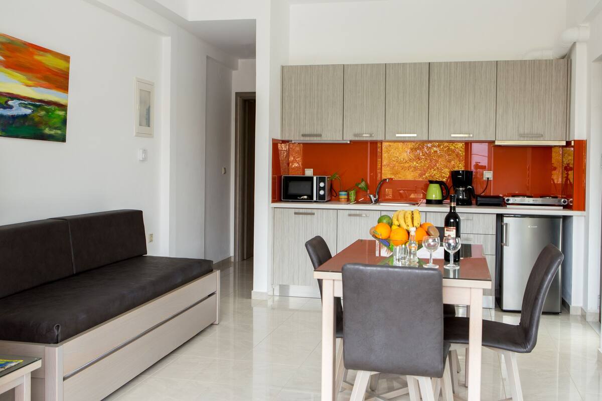 A modern living room with a sofa, dining table, and chairs. A kitchen with appliances and a colorful backsplash is visible in the background.