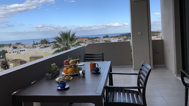 A balcony with a table set for breakfast, a chair, and a view of the sea and buildings. - Angela (Chania)