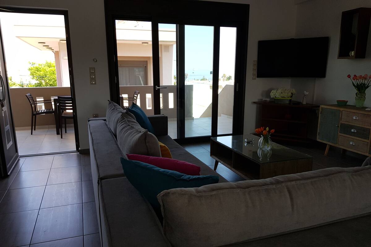 A living room with a sofa, a coffee table, and a TV. There is a balcony with a table and chairs outside.