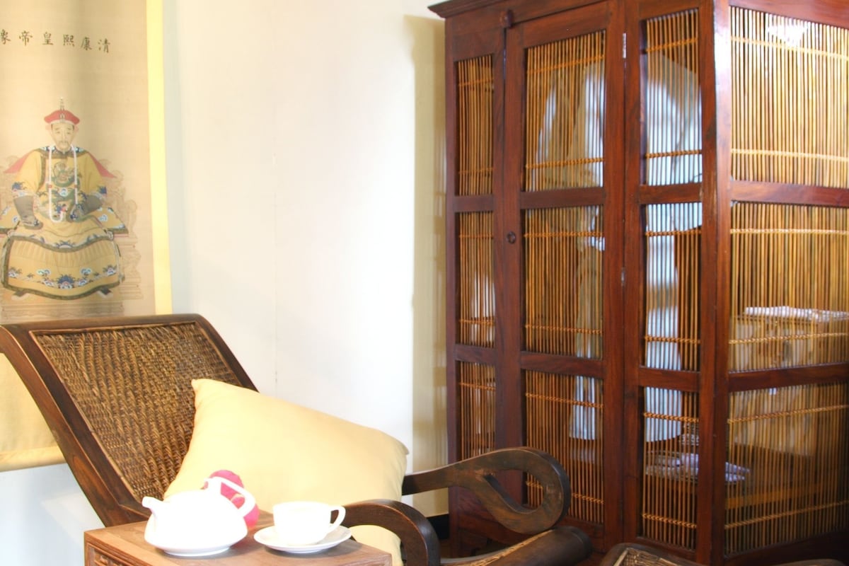 A traditional wooden cabinet with a woven screen door, a wicker chair with a cushion, and a small table with a teapot and cups.