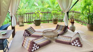 An outdoor seating area with cushioned benches and a woven table, surrounded by lush greenery and a wooden fence. - Ban Keaw Villas (Koh Samui)