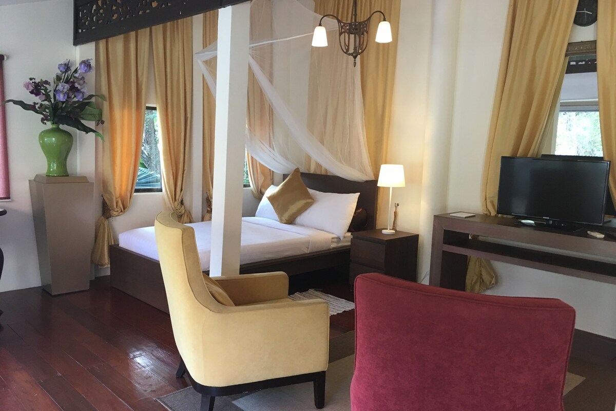 Una habitación de hotel con cama con dosel, un televisor, un sillón rojo y un jarrón verde con flores.