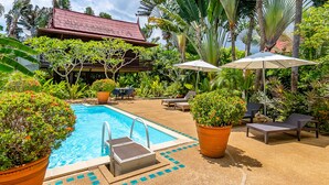 A pool area with lounge chairs and umbrellas, surrounded by potted plants and a building with a red roof. - Ban Keaw Villas (Koh Samui)