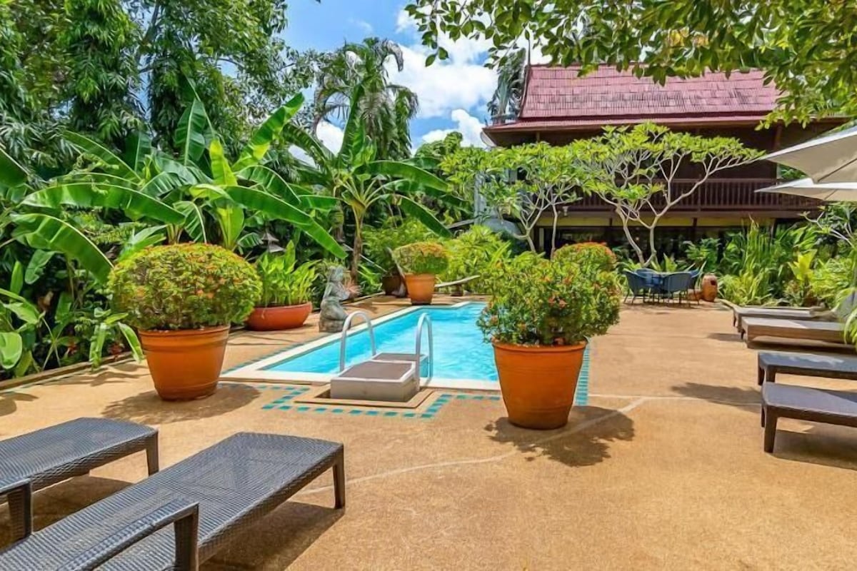 Zona de piscina con sillones de descanso, plantas en macetas y un edificio con techo rojo al fondo.