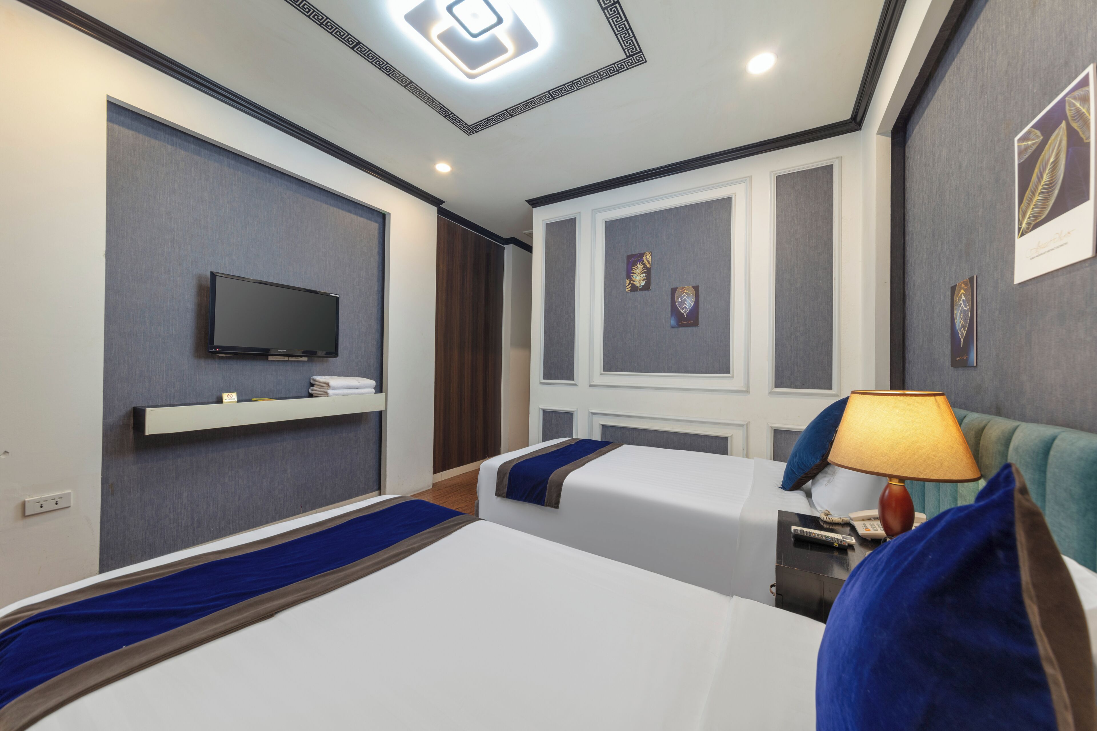 A hotel room with two beds, a TV, a bedside table with a lamp, and artwork on the walls.