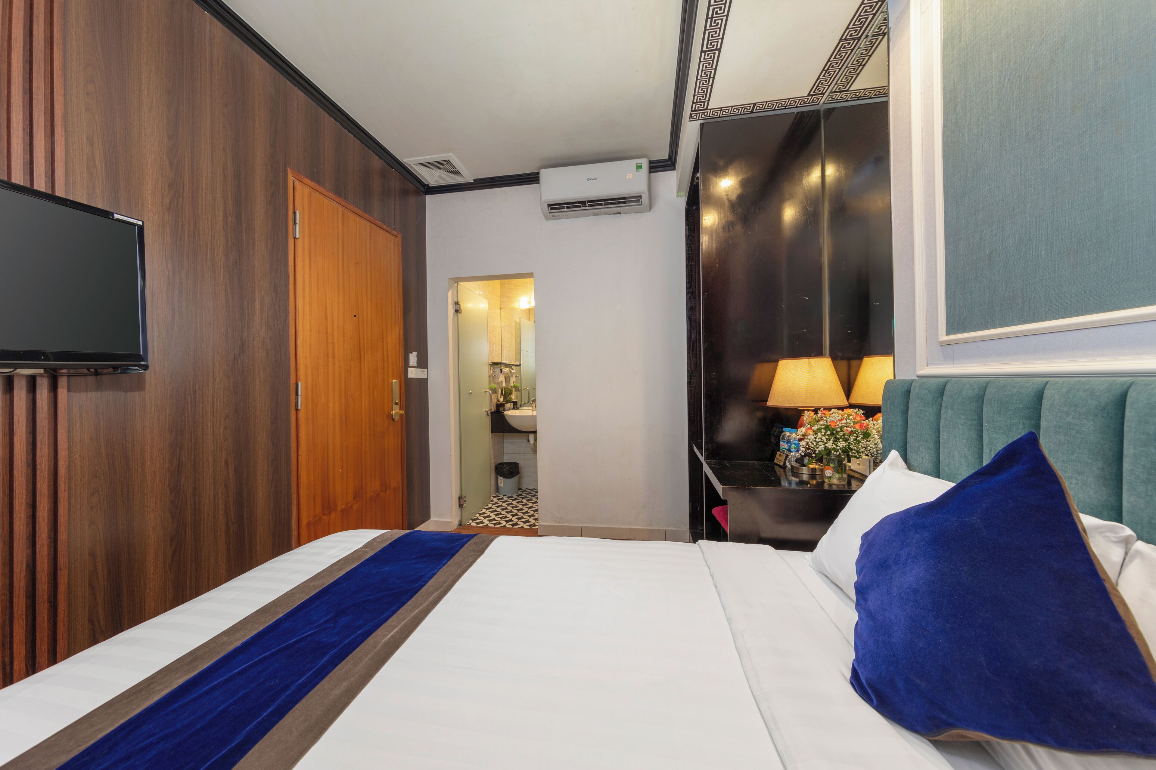 A hotel room with a bed, a television, a wooden wall, a bathroom visible through an open door, and a nightstand with a lamp.
