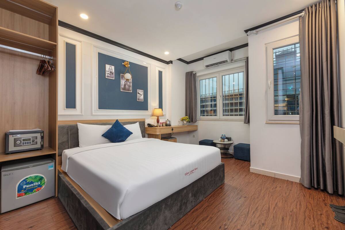 Una habitación de hotel con cama, escritorio, silla, mesita, armario y ventana.