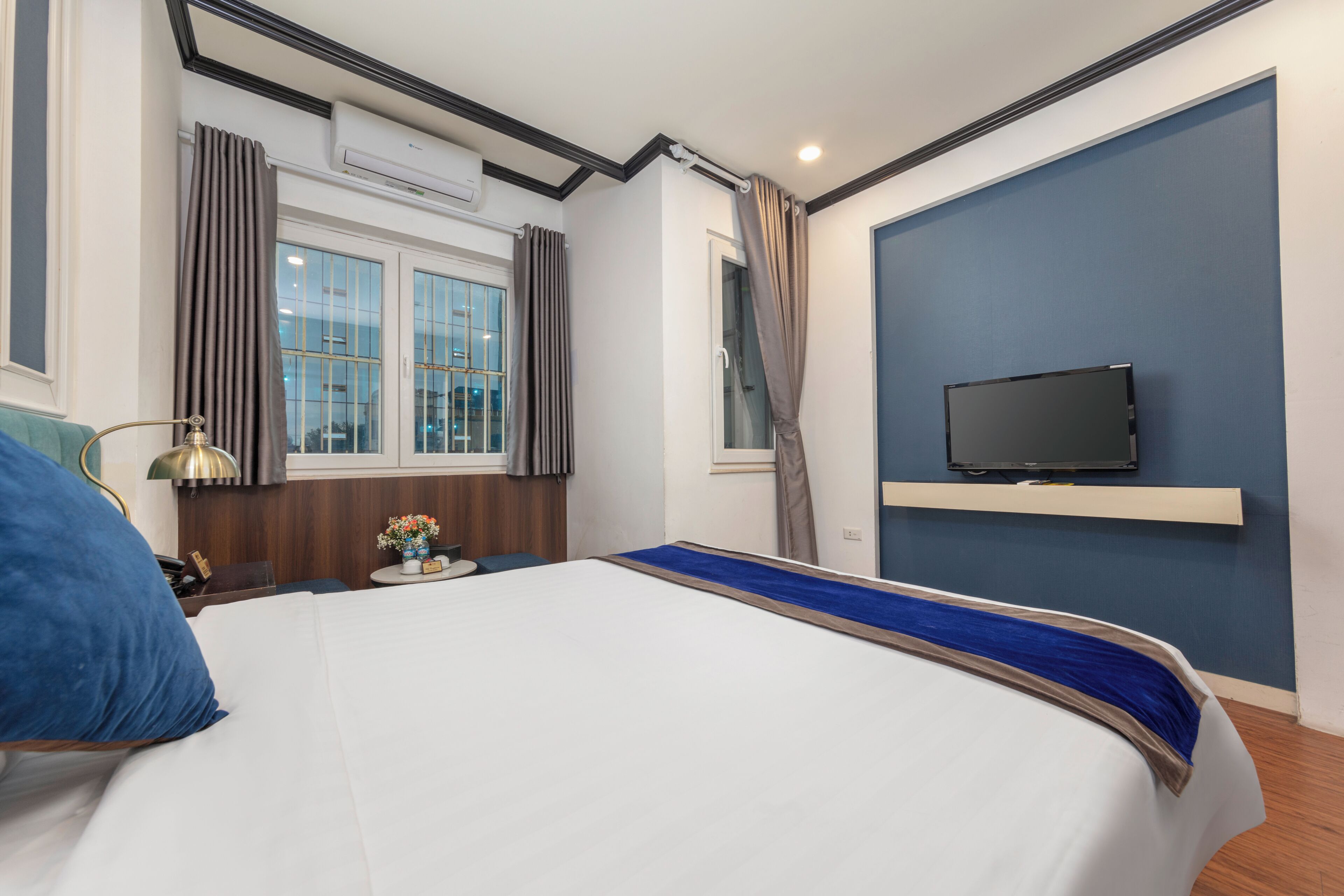 Une chambre d’hôtel avec un lit, un téléviseur fixé au mur, une fenêtre avec des rideaux et une petite table sur laquelle se trouvent une lampe et des fleurs.