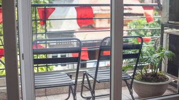 Une fenêtre donnant sur un balcon, où l’on aperçoit des chaises en métal et un pot de fleurs.