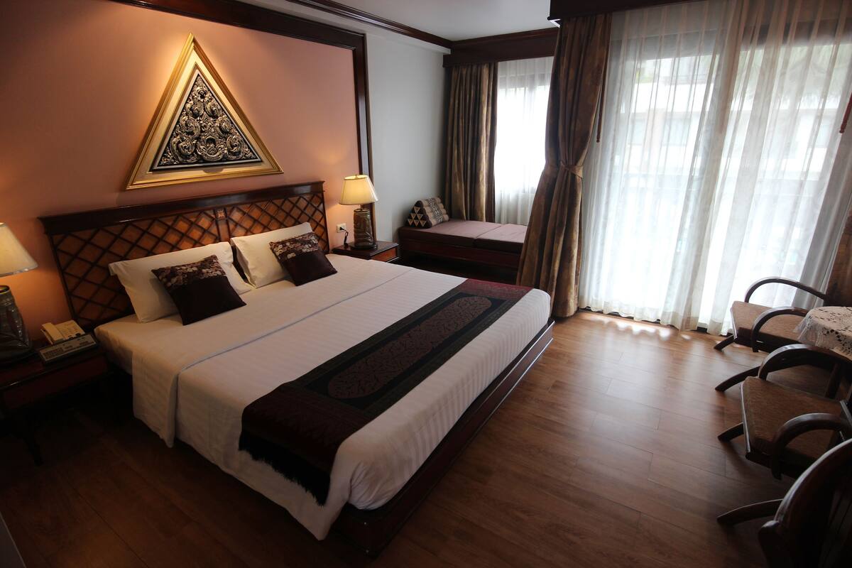 A hotel room with a large bed, a bedside table with a lamp, a decorative wall piece, a window with curtains, and a seating area with chairs.