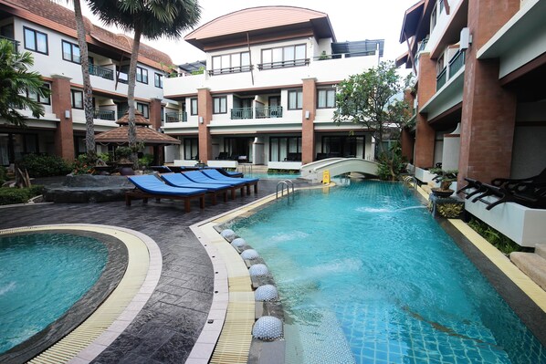 A hotel pool area with multiple lounge chairs, a small bridge, and a surrounding garden.