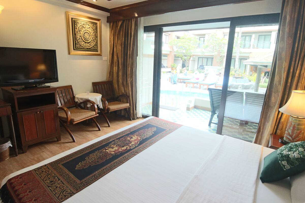 A hotel room with a large bed, a television, a wooden cabinet, a chair, a small table, and a view of an outdoor pool area.
